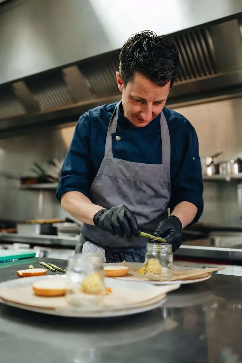 Een chef-kok in een professionele keuken die met zorg een asperge in een weckpotje plaatst bij het bereiden van gerechten.